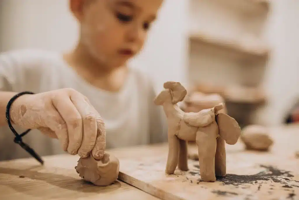 Imagem de uma criança modelando argila para criar uma escultura de um animal, com as mãos cobertas de argila e um sorriso no rosto.
