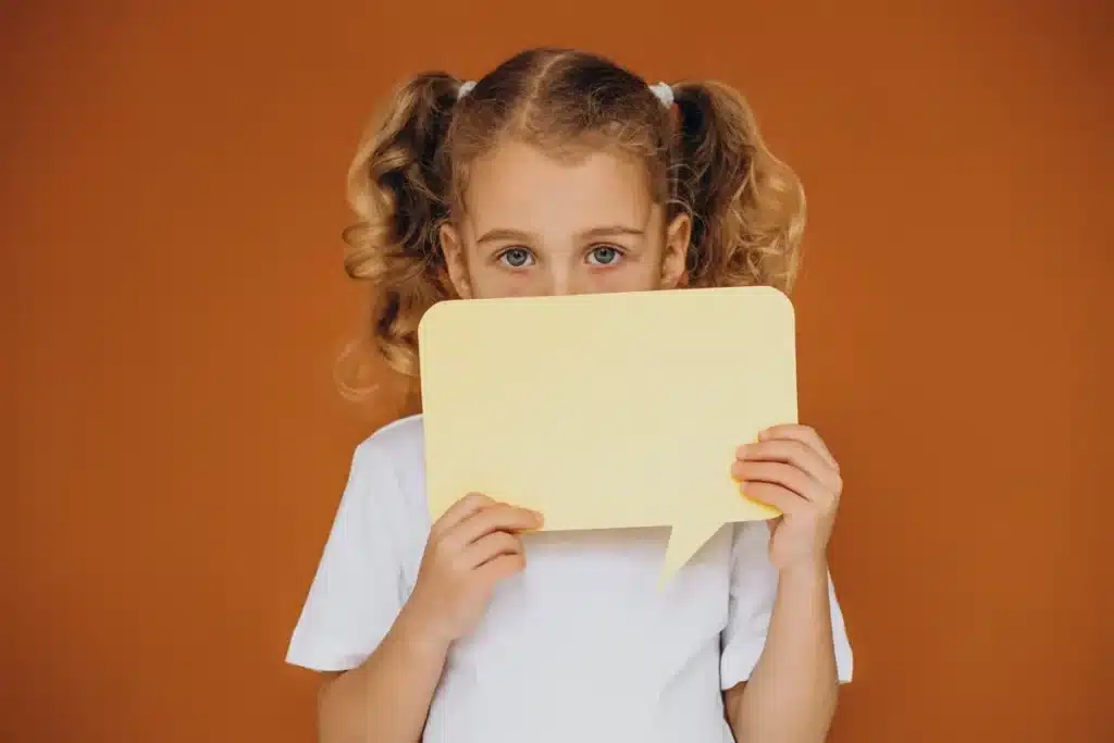 Menina com cabelos cacheados segurando uma placa amarela em forma de balão de fala, com expressão neutra, em fundo laranja.