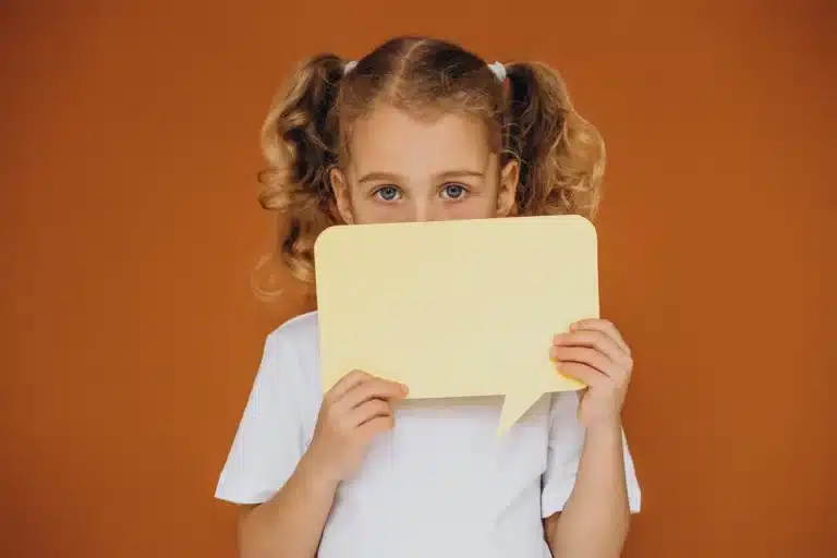 Menina com cabelos cacheados segurando uma placa amarela em forma de balão de fala, com expressão neutra, em fundo laranja.