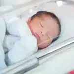 Bebê prematuro dormindo em uma incubadora, envolvido em cobertor, com cuidados de saúde ao fundo