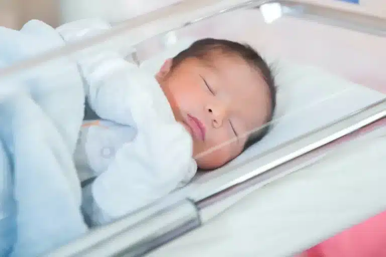 Bebê prematuro dormindo em uma incubadora, envolvido em cobertor, com cuidados de saúde ao fundo