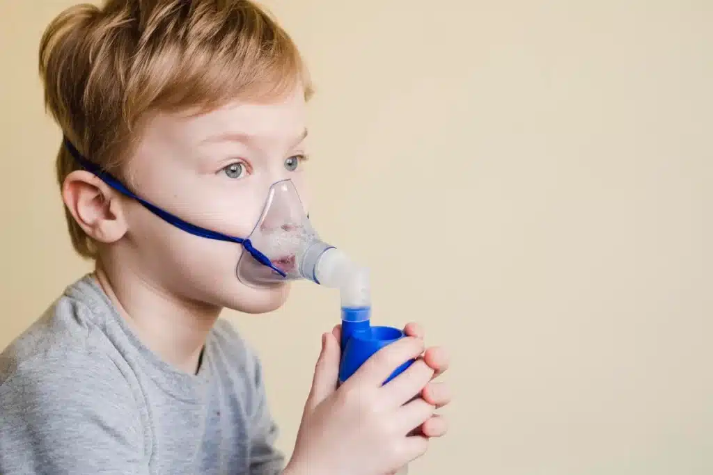 Menino loiro usando máscara de inalação com nebulizador para tratamento respiratório, com fundo bege.