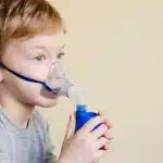 Menino loiro usando máscara de inalação com nebulizador para tratamento respiratório, com fundo bege.