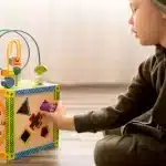 Criança brincando com uma caixa sensorial, colocando uma peça colorida em um dos compartimentos da caixa.