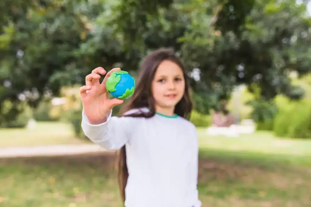 Criança sorrindo e segurando um globo terrestre feito de massa de modelar, com cores vibrantes representando o planeta, em um ambiente ao ar livre, em um jardim ou parque.