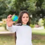Criança sorrindo e segurando um globo terrestre feito de massa de modelar, com cores vibrantes representando o planeta, em um ambiente ao ar livre, em um jardim ou parque.