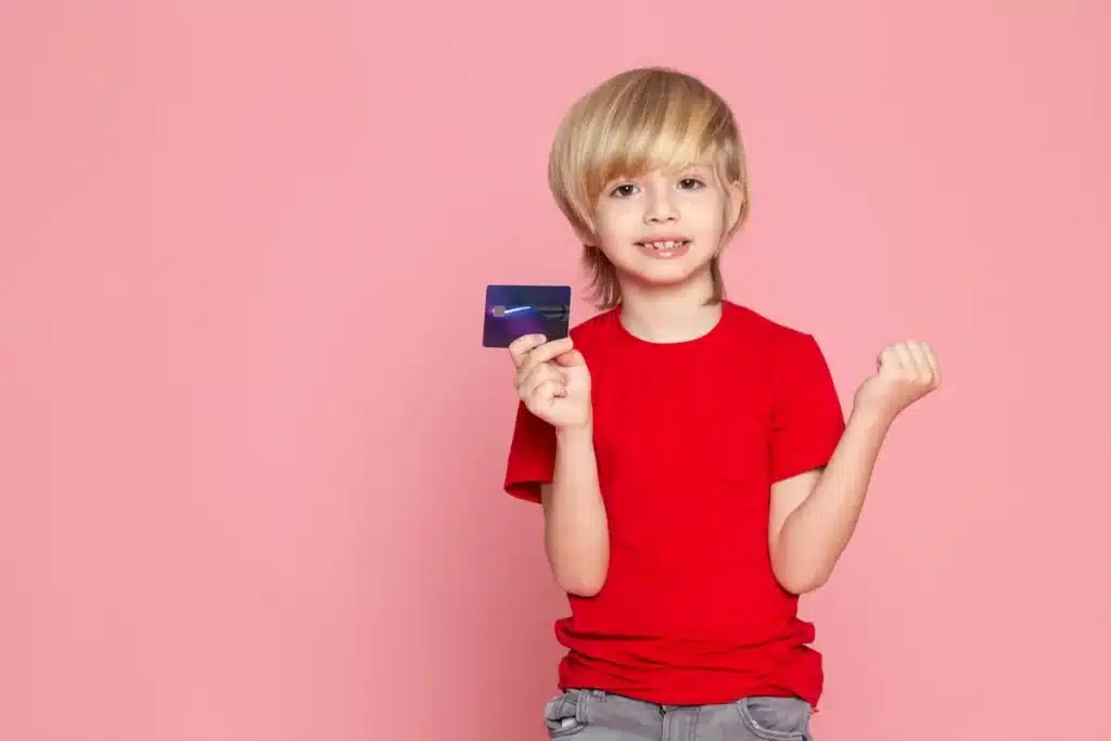 Menino sorrindo e segurando um cartão de crédito, com o braço direito levantado em um gesto de celebração, fundo rosa