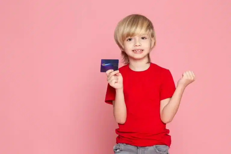 Menino sorrindo e segurando um cartão de crédito, com o braço direito levantado em um gesto de celebração, fundo rosa