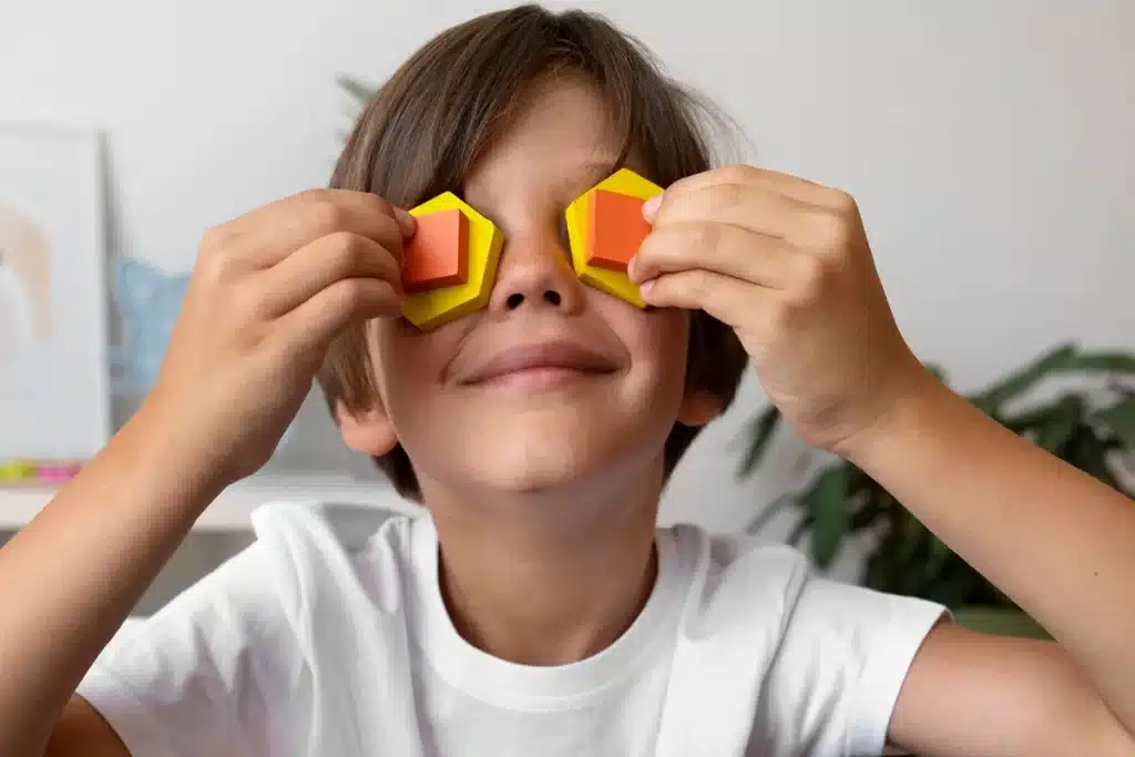 Menino sorridente com camiseta branca cobrindo os olhos com blocos geométricos coloridos, simbolizando criatividade e imaginação infantil.