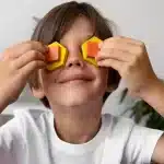 Menino sorridente com camiseta branca cobrindo os olhos com blocos geométricos coloridos, simbolizando criatividade e imaginação infantil.