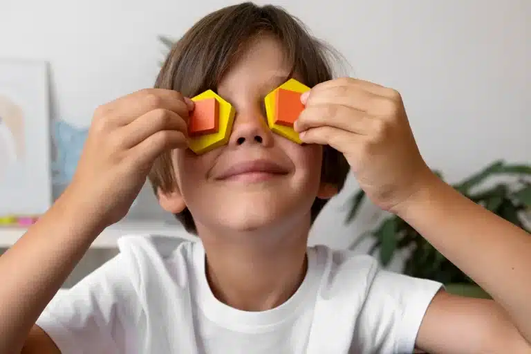 Menino sorridente com camiseta branca cobrindo os olhos com blocos geométricos coloridos, simbolizando criatividade e imaginação infantil.