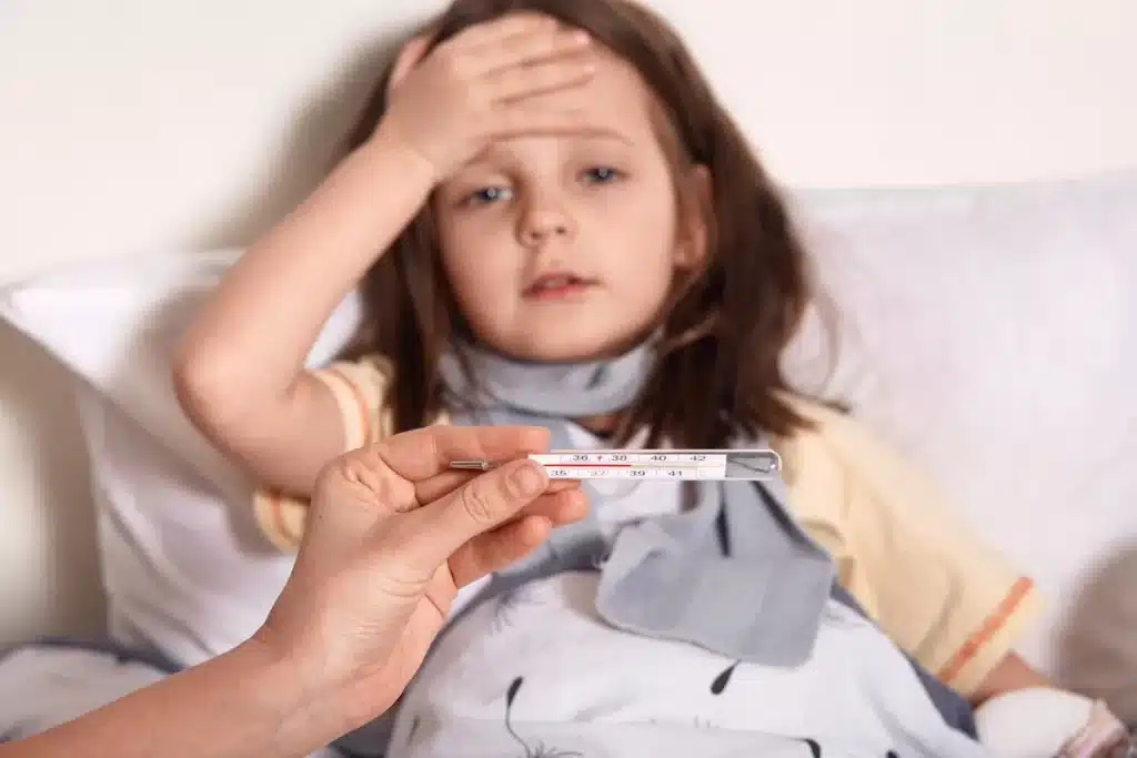 Menina doente deitada na cama com expressão de mal-estar, mão na testa e termômetro sendo segurado em primeiro plano.