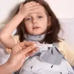 Menina doente deitada na cama com expressão de mal-estar, mão na testa e termômetro sendo segurado em primeiro plano.