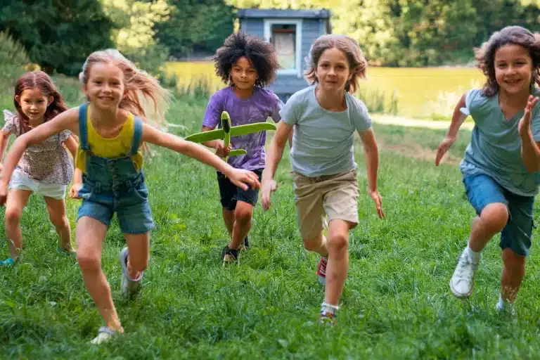 Imagem de cinco crianças correndo e se divertindo ao ar livre durante uma atividade de gincana, com um avião de brinquedo em uma das mãos