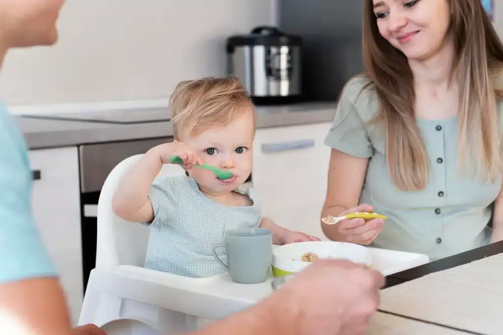 Bebê usando uma colher verde para se alimentar enquanto a mãe o observa, em uma cozinha moderna e tranquila.