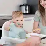 Bebê usando uma colher verde para se alimentar enquanto a mãe o observa, em uma cozinha moderna e tranquila.