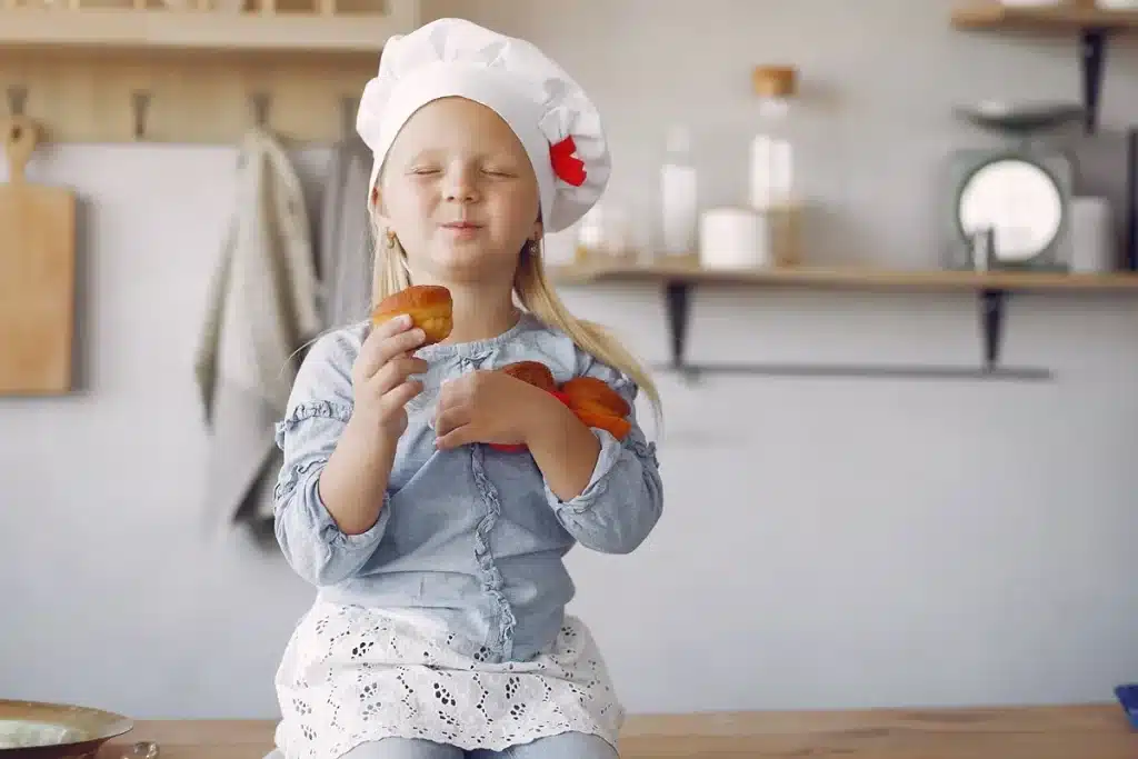 Garotinha com chapéu de chef segurando bolinhos caseiros, sorrindo de felicidade na cozinha