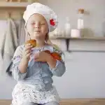 Garotinha com chapéu de chef segurando bolinhos caseiros, sorrindo de felicidade na cozinha