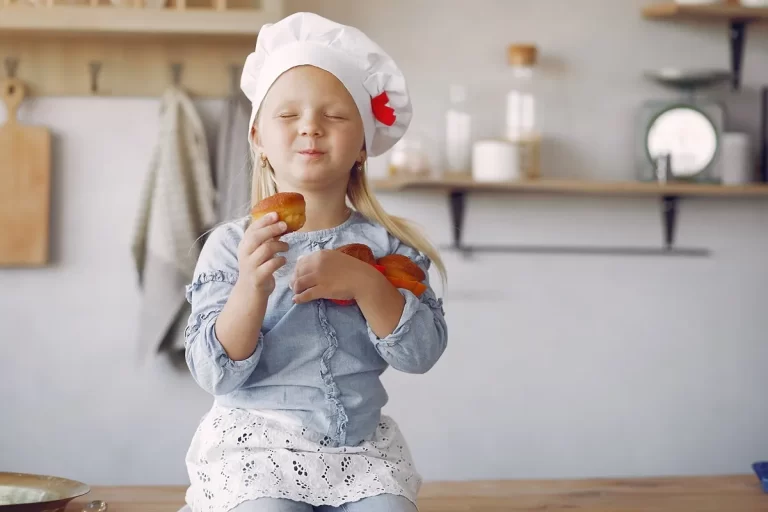 Garotinha com chapéu de chef segurando bolinhos caseiros, sorrindo de felicidade na cozinha