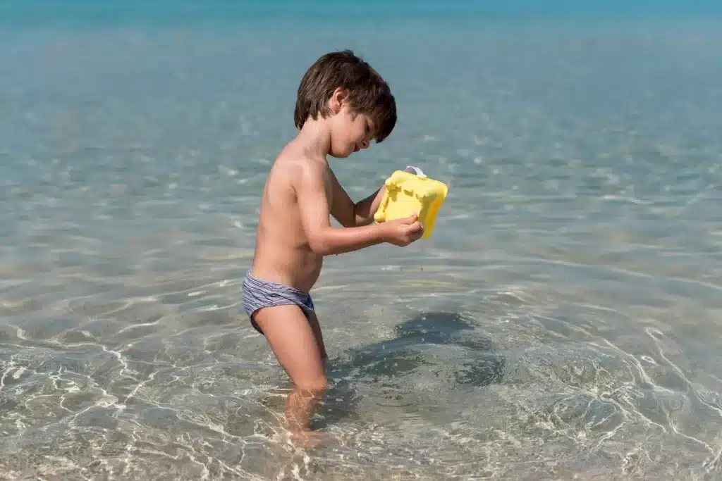 Criança de sunga listrada brincando na água da praia, segurando um brinquedo amarelo. O cenário é claro e tranquilo, com águas cristalinas e céu azul