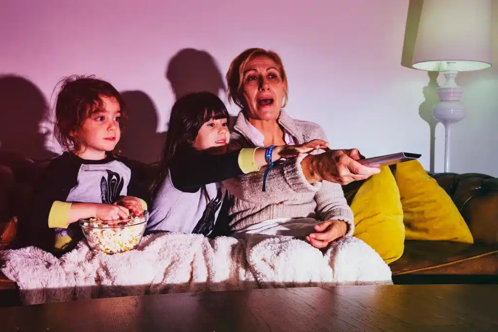 Mulher e duas crianças assistindo a uma série infantil, compartilhando um momento de diversão no sofá com pipoca.