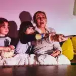 Mulher e duas crianças assistindo a uma série infantil, compartilhando um momento de diversão no sofá com pipoca.