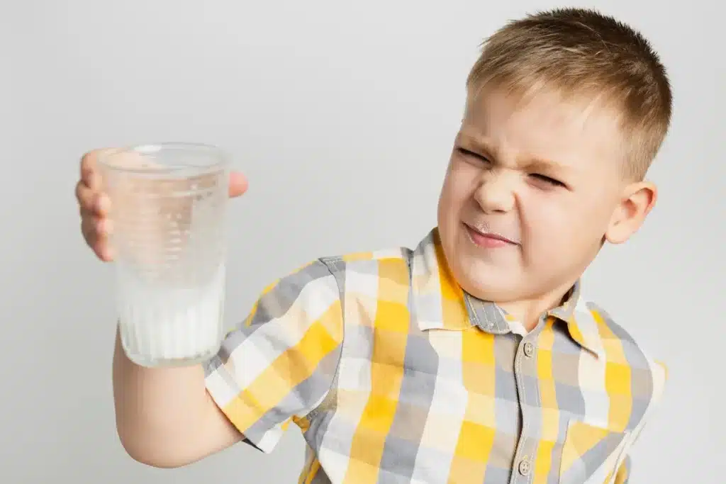 Criança segurando um copo de leite com uma expressão de desgosto, possivelmente indicando uma reação a sintomas de intolerância à lactose, como cólicas ou desconforto abdominal.
