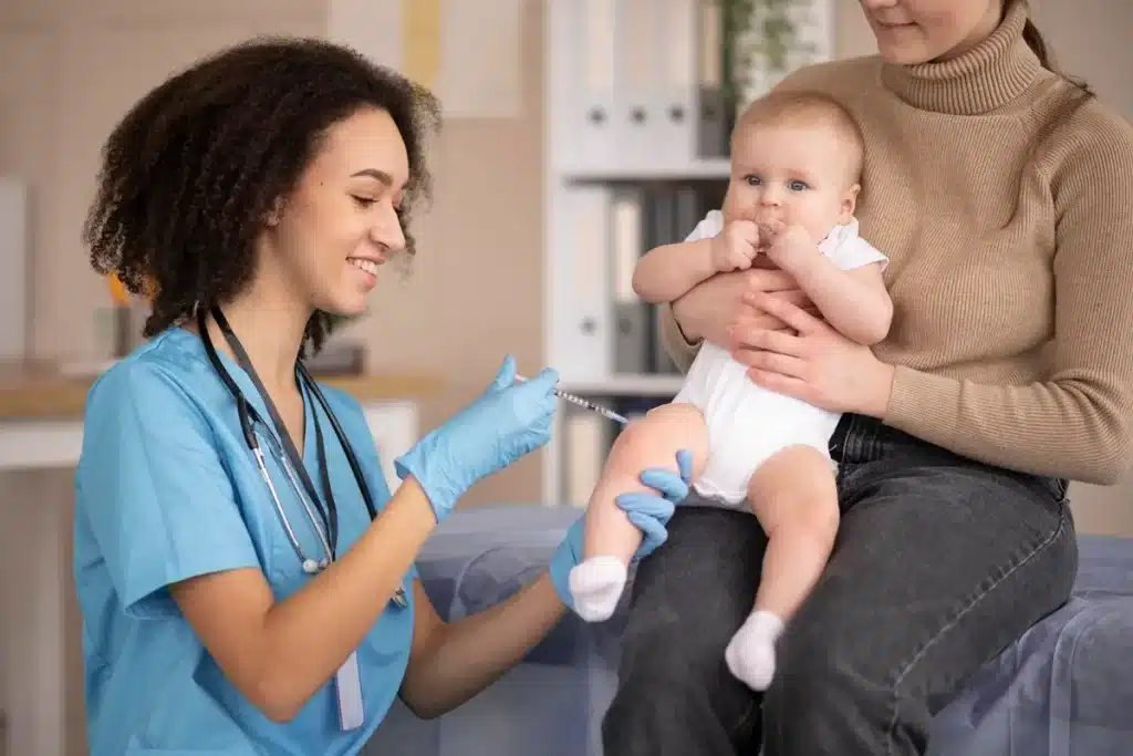 Enfermeira aplicando vacina na coxa de um bebê no colo da mãe em consultório pediátrico.