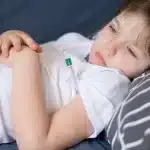 Criança deitada em cama com camiseta branca, segurando o termômetro, mostrando sinais de febre e cansaço.