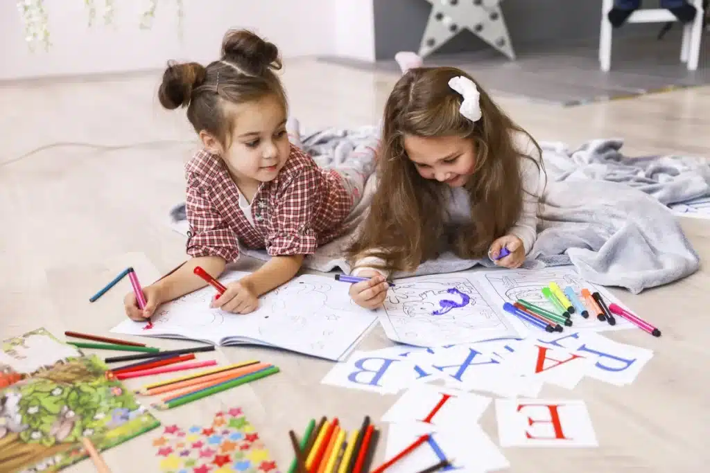 Duas meninas desenhando e colorindo com lápis de cor e canetinhas, rodeadas por letras do alfabeto em cartões. Atividade educativa e criativa para crianças.