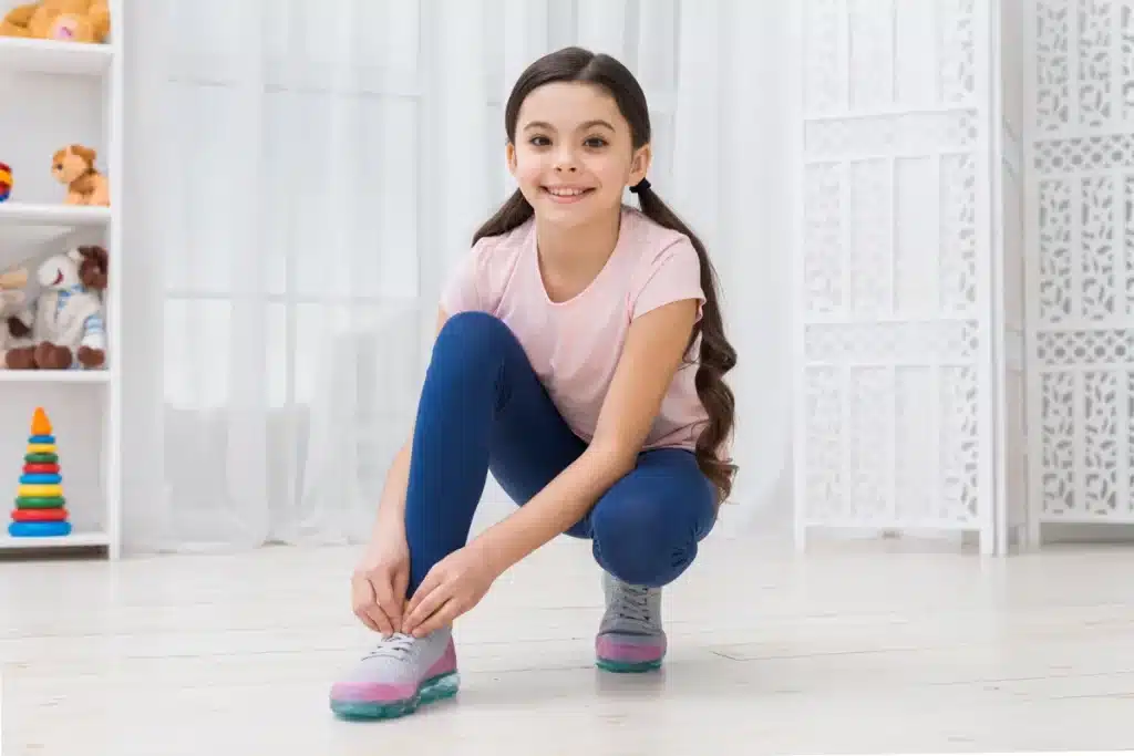 Menina sorridente amarrando os cadarços de seu tênis infantil rosa e cinza em um quarto decorado com brinquedos.