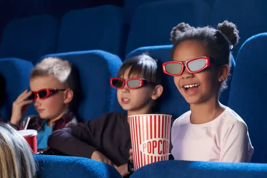 Imagem de crianças assistindo a um filme em 3D em um cinema, usando óculos 3D e segurando pipoca, com um sorriso no rosto.