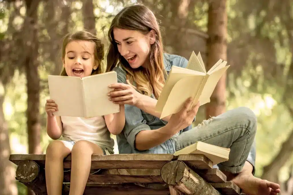 Mãe adulta e menina sentadas em banco de madeira lendo livros sob árvores em ambiente natural
