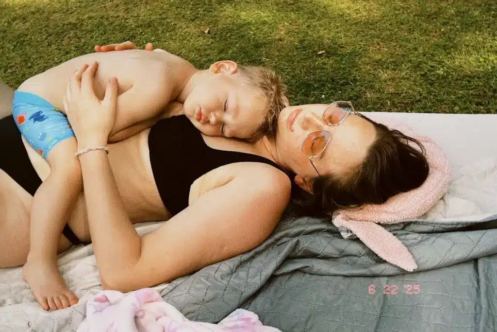 Mulher de óculos de sol e biquíni deitada em um cobertor na grama, com um menino pequeno dormindo aninhado em seus braços, transmitindo afeto e segurança.