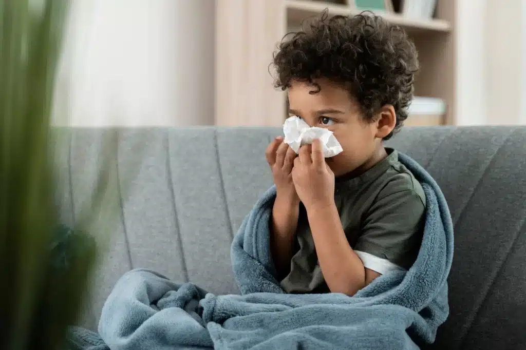Criança com rinite alérgica usando lenço de papel no nariz, sentada no sofá e coberta com manta azul — sintomas comuns de alergia respiratória infantil.