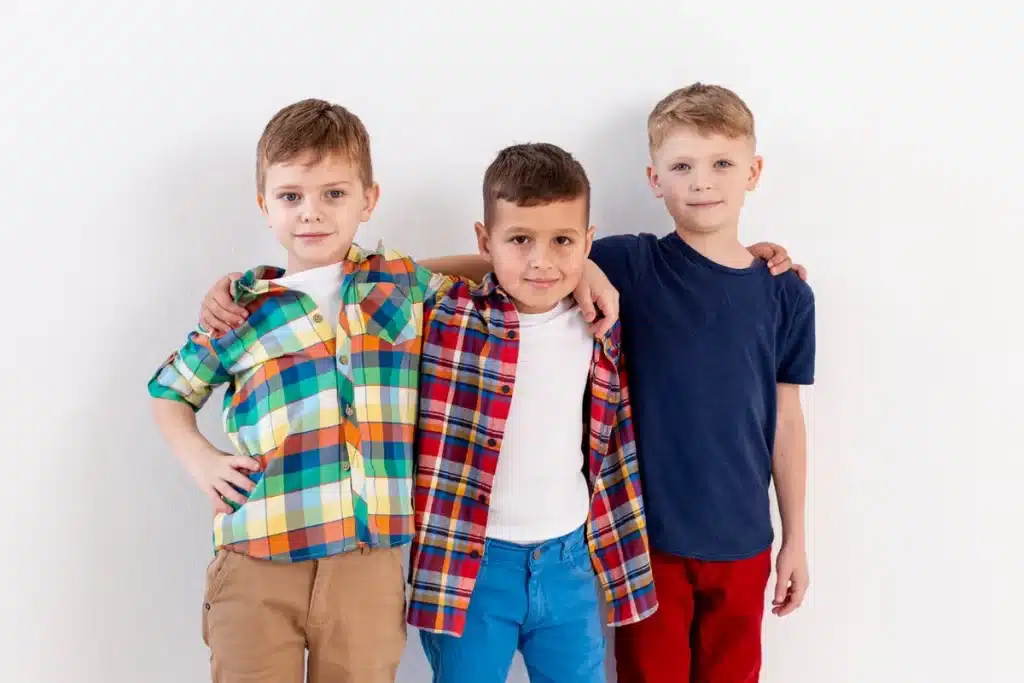 Três meninos com roupas infantis modernas e coloridas: dois com camisas xadrez e camisetas brancas, e um com camiseta azul-marinho e calça vermelha, posando juntos em frente a parede branca
