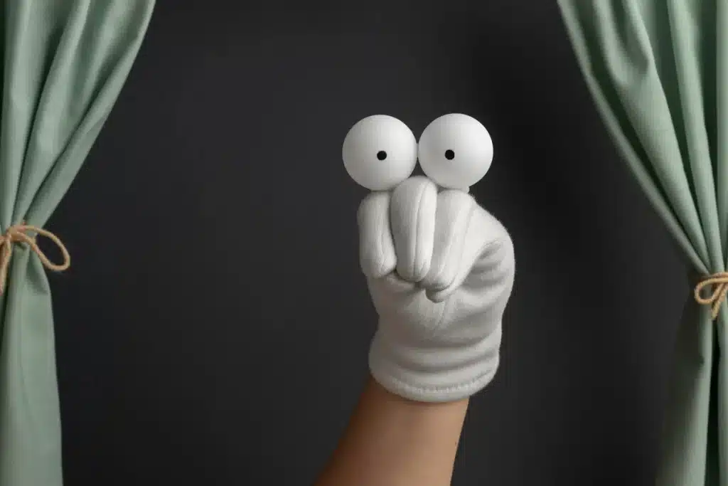 Imagem de uma mão com um fantoche improvisado feito de bolas de pingue-pongue, com cortinas de teatro ao fundo.