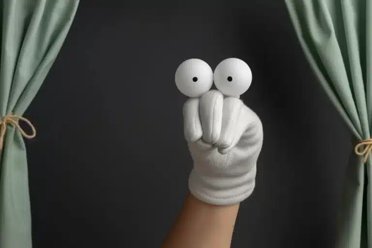 Imagem de uma mão com um fantoche improvisado feito de bolas de pingue-pongue, com cortinas de teatro ao fundo.