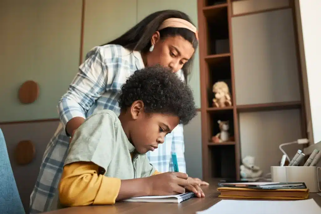 Imagem de uma mãe apoiando seu filho enquanto ele escreve no caderno, ambos concentrados no estudo. A mãe observa atentamente, oferecendo apoio e orientação.