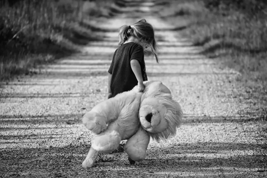 Fotografia em preto e branco de uma menina caminhando sozinha em uma estrada rural, carregando um urso de pelúcia, evocando sentimentos de isolamento e possível impacto do abuso psicológico.