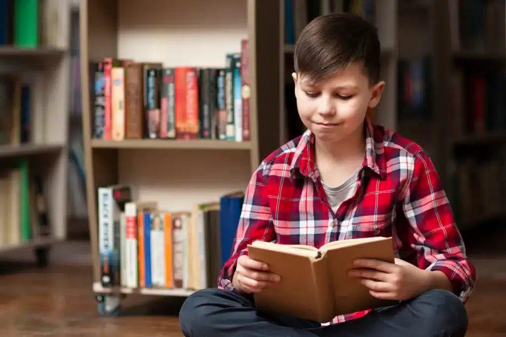 Menino sentado no chão em uma biblioteca, vestindo camisa xadrez vermelha, lendo atentamente um livro