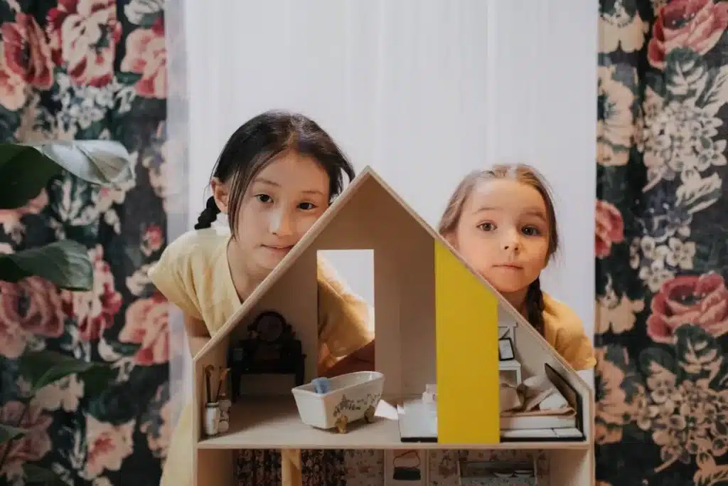 Duas meninas curiosas espiando por trás de uma casa de bonecas de madeira com miniaturas de móveis. A imagem destaca "brinquedos de meninas" que promovem a criatividade e a narrativa.