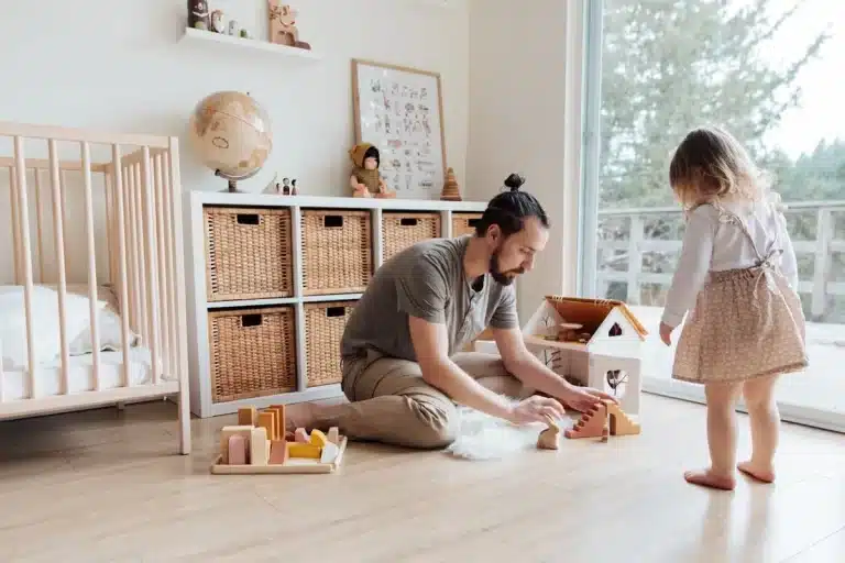 Pai e filha brincando juntos no chão do quarto infantil com uma casa de bonecas de madeira, blocos de montar e outros "brinquedos educativos".