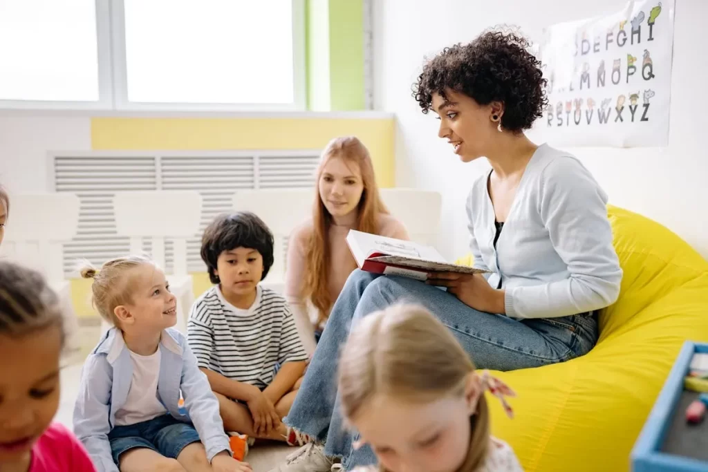 Professora sentada em puff amarelo lendo um livro para grupo de crianças atentas no chão, em um cantinho de leitura acolhedor em sala de aula no Brasil.