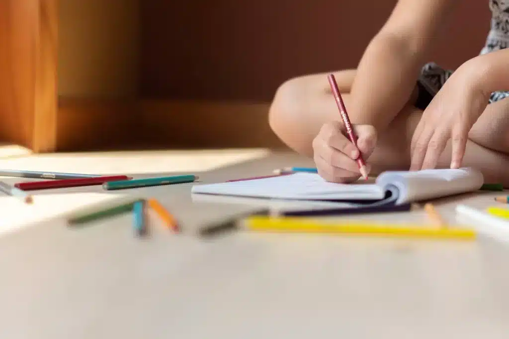 Mãos de criança segurando um lápis de cor vermelho e escrevendo ou desenhando em um caderno aberto no chão, rodeado por outros lápis coloridos. Ilustra a prática de escrita e "como escrever o nome" de forma lúdica.