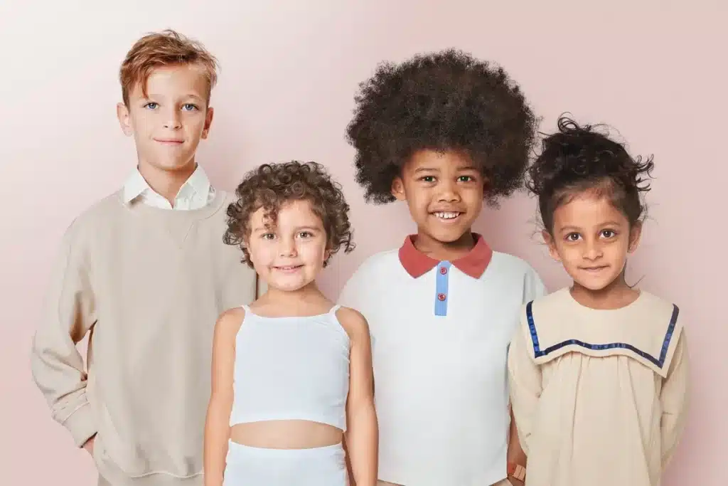 Quatro crianças sorrindo em frente a fundo claro: um menino loiro de suéter bege, uma menina de cabelo cacheado curto com top branco, uma menina negra de cabelo afro usando camisa polo clara e uma menina de pele morena com vestido bege de gola quadrada