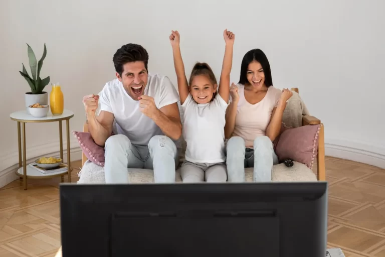 Família (pai, filha e mãe) sentada no sofá comemorando e assistindo televisão, representando a alegria de ver desenhos infantis juntos.