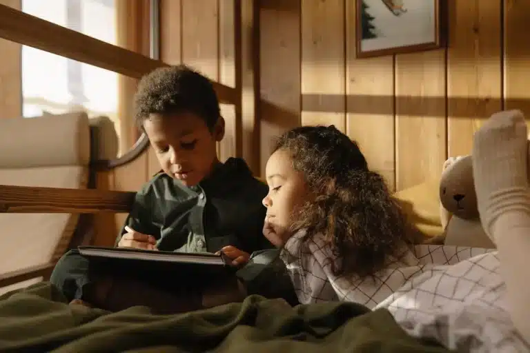 Duas crianças em cama ou beliche, menino desenhando em tablet, menina observando, atividade de escrita criativa, aprendizado e imaginação infantil.