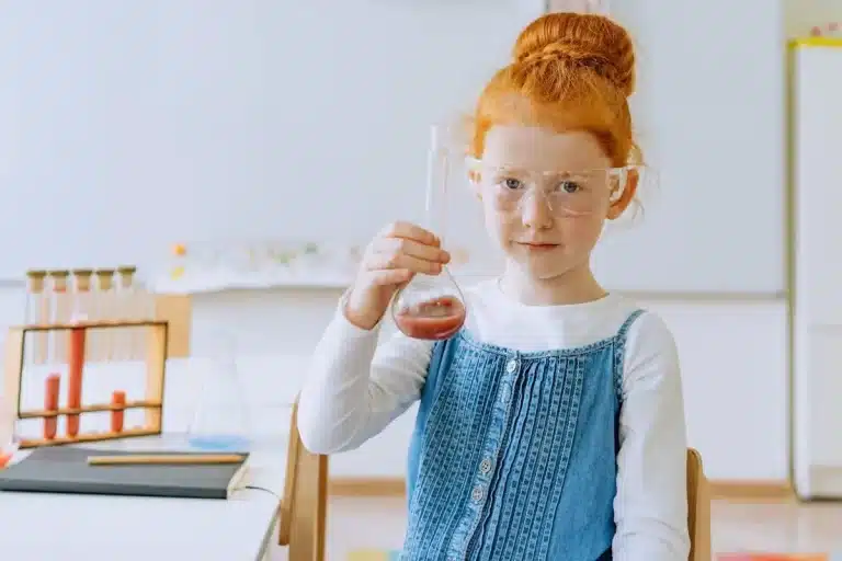 Jovem garota ruiva usando óculos de proteção e segurando um balão de laboratório com líquido rosa. Experimento científico infantil.