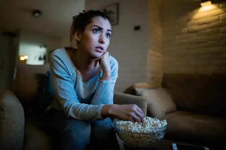 Mulher sentada em sofá segurando tigela de pipoca, com expressão emocionada e olhar fixo na tela do filme